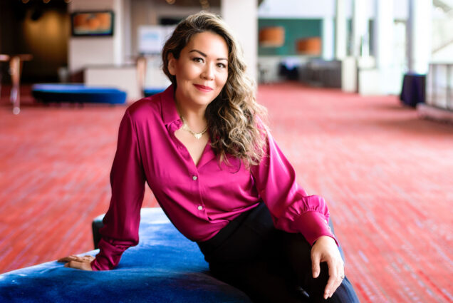 Boston University alum Nina Yoshida Nelsen wearing a fuschia blouse and sitting on a blue sofa. She has been named Artistic Director of Boston Lyric Opera.