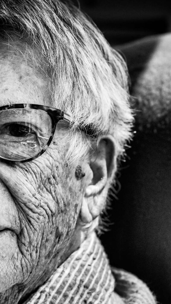 Photo: an up-close portrait of a centenarian wearing glasses and a sweater. Photo is black and white