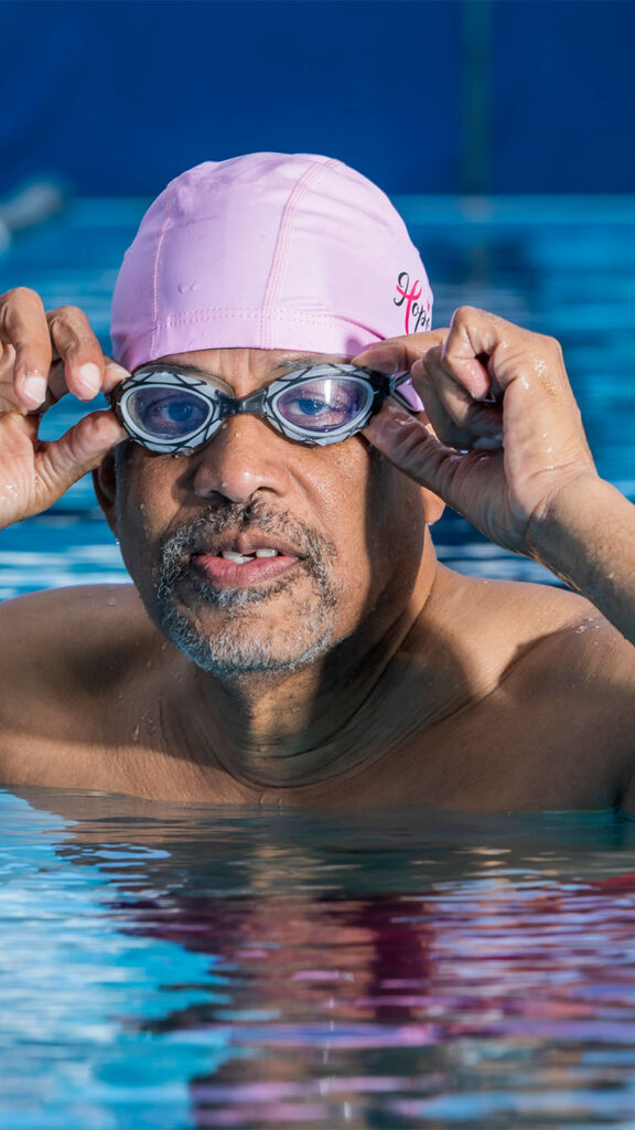 Photo: An older black man wearing goggles and a swim cap poses for a portrait in a pool