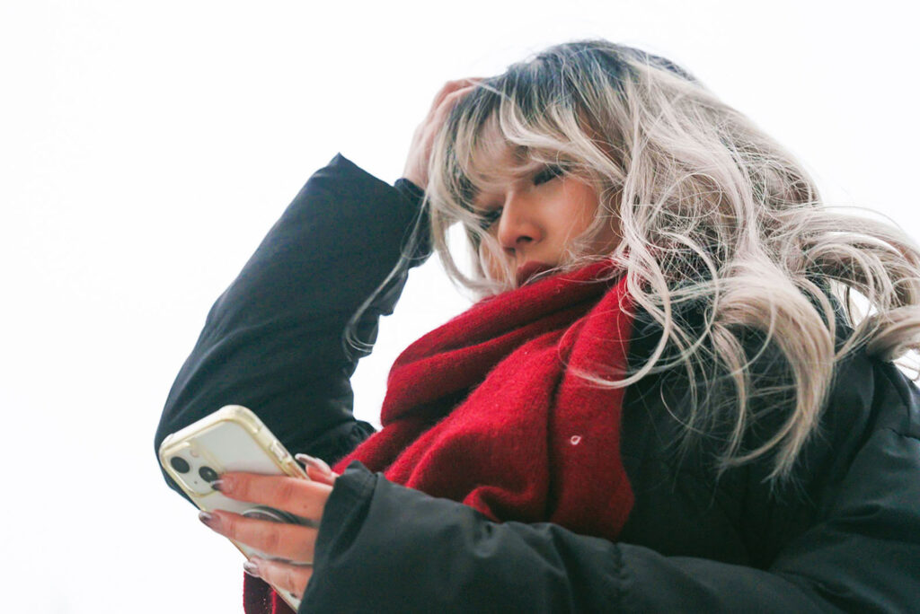 Photo: A young woman holds her head in her hand as she looks down at her phone. She has dyed blonde hair and wears a read scarf around her neck.