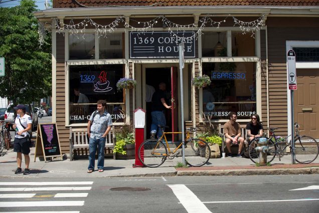 Photo of the facade of 1369 Coffee House in Inman square. A Black and white sign hangs over the door. People sit on the benches outside chatting, while others wait to cross the street. Several bikes are parked out front, and neon signs that read "1369," "Espresso," and a coffee cup with red steam hang in the window.