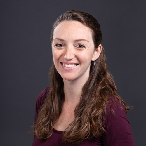 Photo of Amy Laskowski. A white woman with long brown hair pulled into a half up, half down style and wearing a burgundy top, smiles and poses in front of a dark grey backdrop.