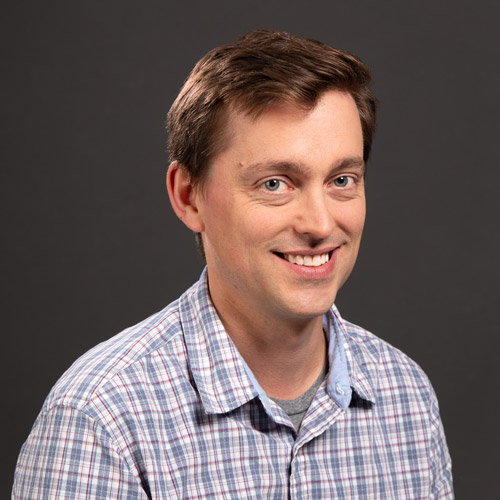 Photo: Headshot of Marc Chalufour. A white man with brown hair and wearing a red and blue plaid shirt, smiles and poses in front of a dark grey background.