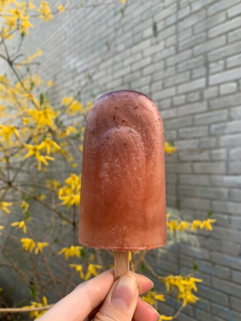 Photo of a popsicle in hand; the popsicle is light red and pink. In the background a spring tree with yellow flowers is seen.