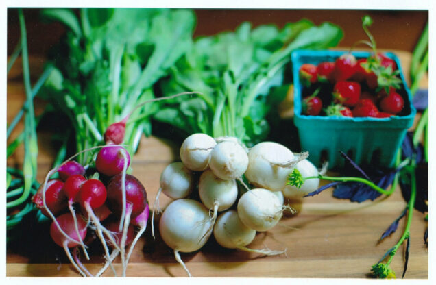 Image of white and red radishes with greens laid on a table; strawberries are seen in the background.