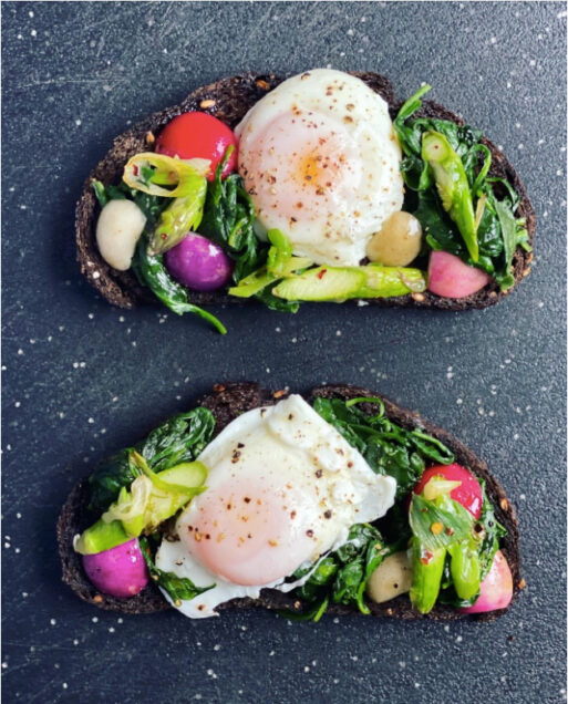Two breakfasts toasts on dark rye or pumpernickel bread side by side on a dark countertop. Each toast has eggs, greens, and tomato slices.
