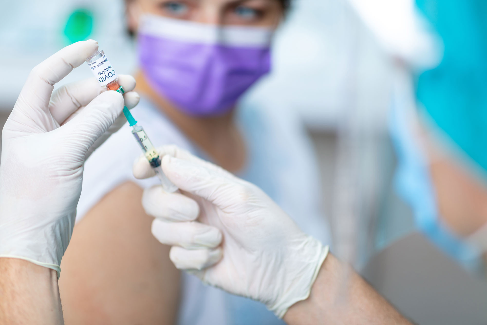 A photo of someone with gloved hands holding a vial that reads "COVID-19 Vaccine" and a needle. A patient is in the background wearing a mask and holding their sleeve up in preparation of receiving the vaccine.