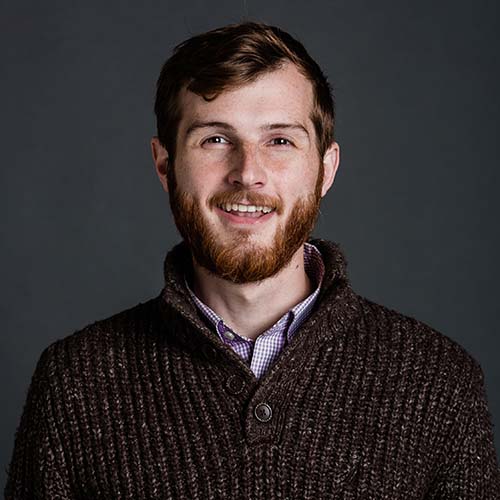 Photo of Andrew Hallock, a young white man with reddish hair and beard. He wears a brownish, gray sweater and smiles.