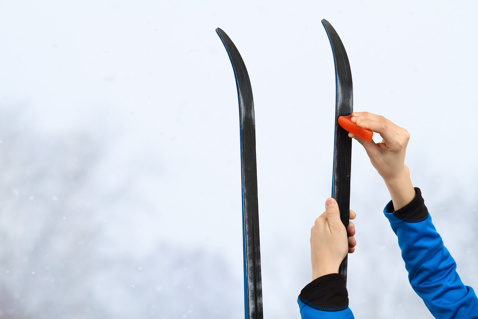 Photo: Closeup of two hands applying wax to black skis. Blurry background shows a snowy scene.