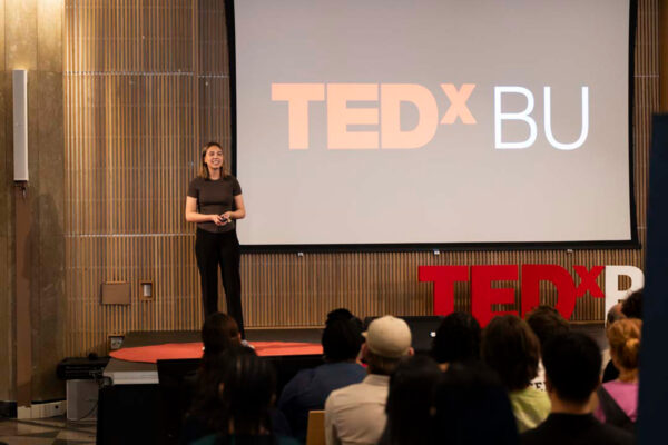Photo: Marie Grochowski (CAS’26) gave a talk titled “What Fungi Can Teach Us About Global Immigration” at the annual TEDxBU conference. A woman stands in front of a large audience and the words "Tedx BU" are on the screen behind