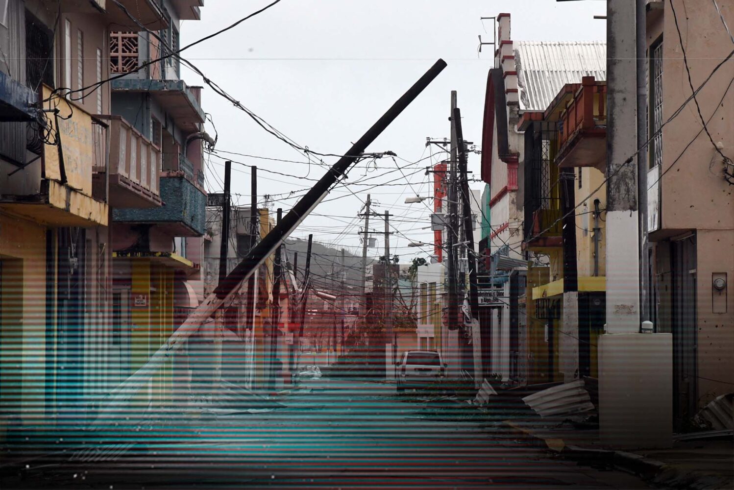 Photo: A picture of a town that has been hit by a hurricane. There is a telephone pole leaning into the street and debris around. There is a glitch overlay on the image
