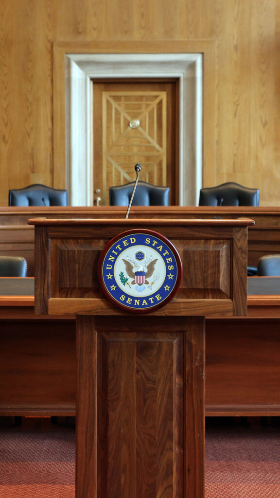 Photo: A picture of a podium with the words "United States Senate" on it. Behind the podium there are tables and chairs