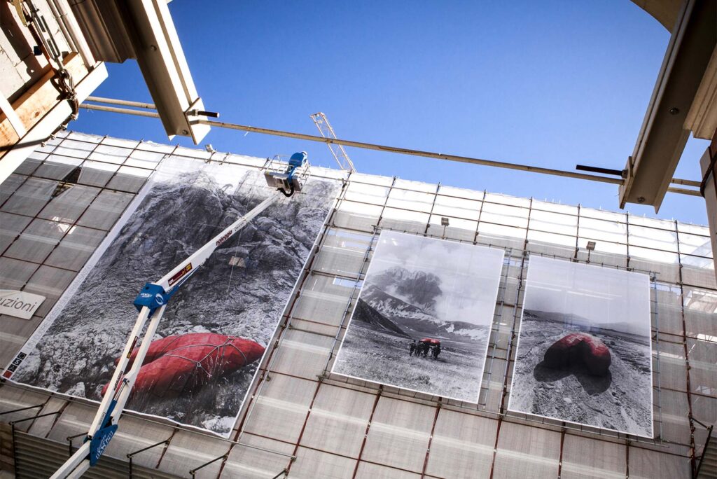 Photo: A man on a scissor lift creating art on the side of a construction site. The art pieces are large black and white landscape paintings with pops of red