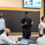 Photo: Three young men stand at the front of a classroom presenting their pitches. On the left, a Black man with a flat top fade wears a blue button up and navy slacks. In the middle, a Black man with his hair pulled into a puff wears a black button up with black slacks. And on the right, a young Asian man with long hair wears a gray polo and slacks.