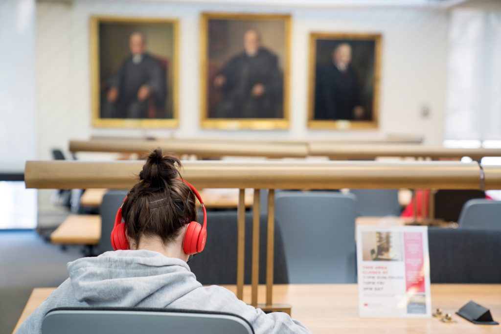 law student sitting with bright red headphones studying in the law library with three portraits of persons in judicial garb