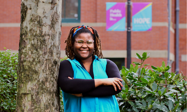 Almira Lewis, wearing a long black top and a bright blue blouse, leans against a tree on campus. A lamp post in the background says "Public Health is... HOPE."