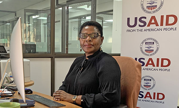 Muka Chikuba-McLeod sits at a desk in front of a USAID sign