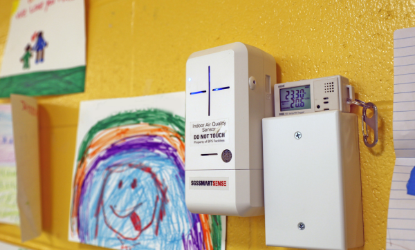 An indoor air quality sensor installed on a yellow wall surround by young children's artwork