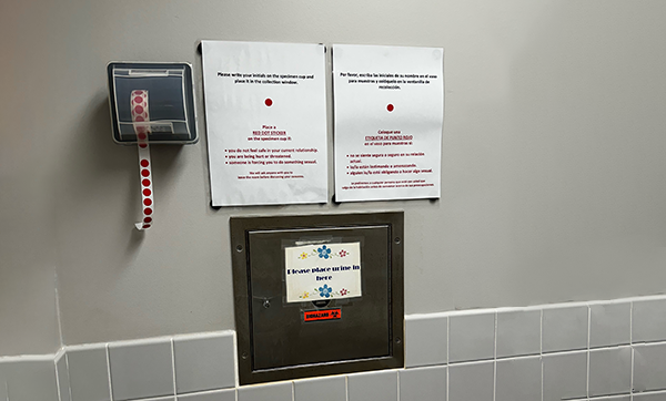 A roll of red circle stickers hangs from a box on a bathroom wall. One sticker is missing.