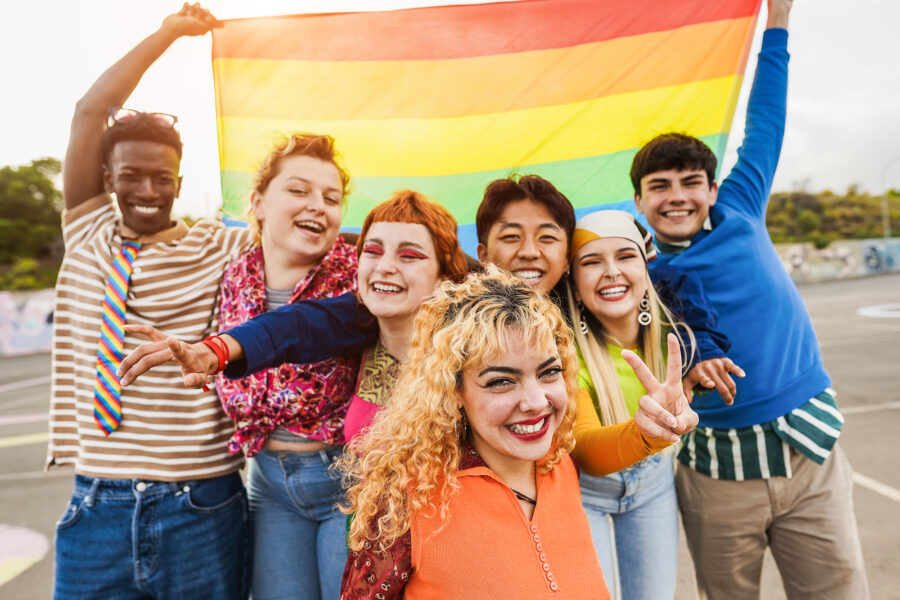 Young diverse people having fun holding LGBT rainbow flag outdoor - Focus on center blond girl