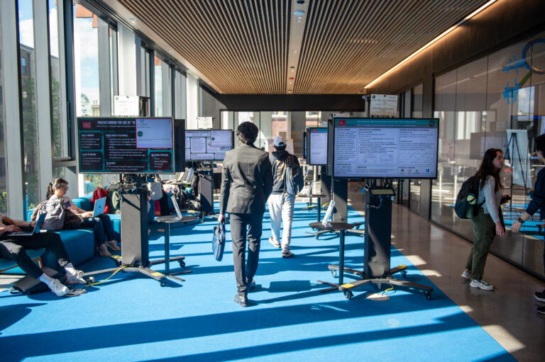 In a spacious room, screens are set up for an expo. Some students can be seen hanging around.