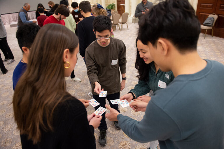 Five students stand in a small circle, engaged in a card game.