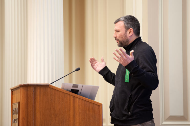 A faculty member, standing on a podium, talks and gestures with his hands.
