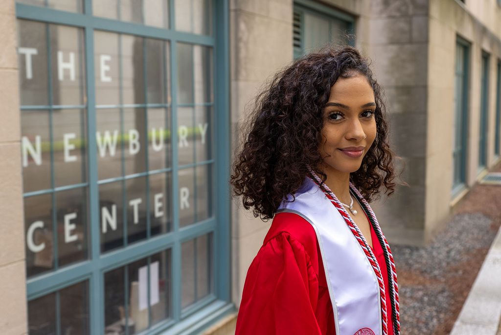 Photo: A young woman with short, curly hair poses in front of BU's Newbury Center in her graduation regalia.
