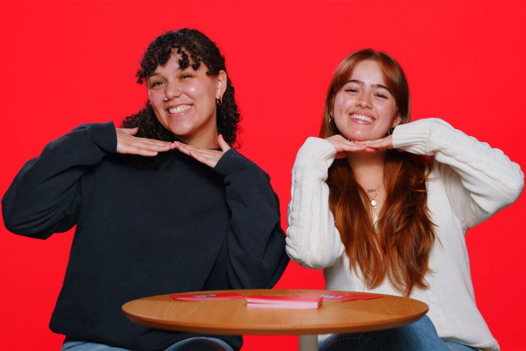 Photo: Two college students in front of a red background smile and hold their hands under their chins