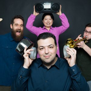 Boston University Ph. D. candidate Reza Ghaffarivardavagh poses with his research team. Reza holds the noise cancellation devices his team developed over his ears while his teammates make noise banging a pot, playing a trumpet, and a boombox in the background