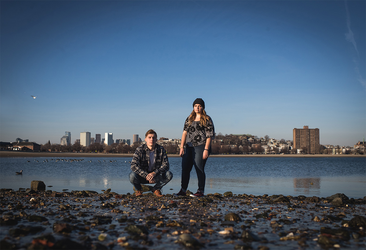 Portrait of Jacob Jaskiel, kneeling, and Hayley Goss, standing, on a Boston Harbor beach with the Boston skyline in the background.