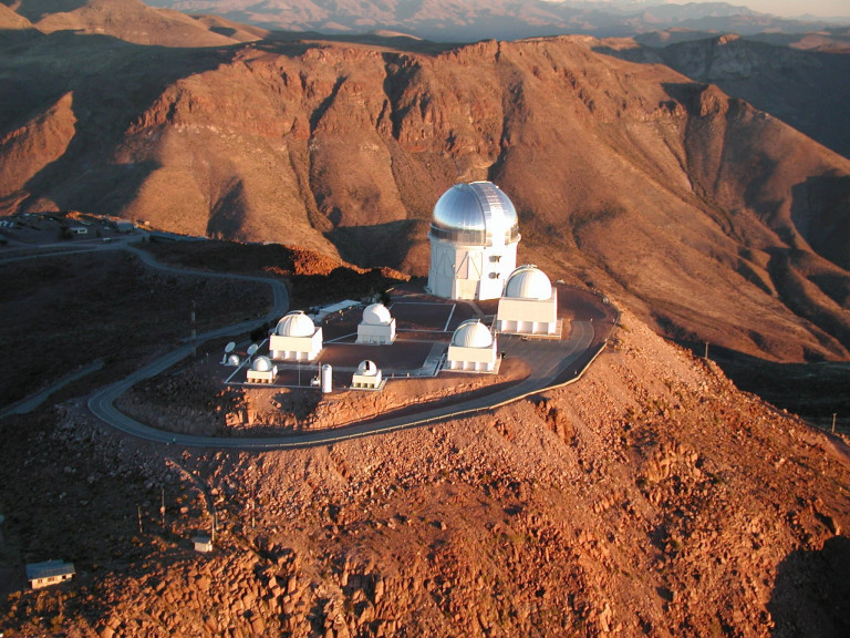 The double door on the ground level of the telescope dome provides a sense of scale (Figure 6). Just inside those doors is a control room from which the camera and telescope are operated each night. Four stories up sits the base of the telescope. The dome is a few stories tall and is covered with aluminum sheets to reflect sunlight during the day. For observations, a large slit, located to the right of the dome, retracts after sunset to allow the telescope and camera to see the night sky. The dome rotates depending on which way the telescope is aimed. The Blanco telescope is probably most famous for its use in the discovery of cosmic acceleration in 1998 by two teams of scientists who studied supernovae in distant galaxies. The 2011 Nobel Prize in physics was awarded to Saul Perlmutter, Adam Riess, and Brian Schmidt for this discovery.
