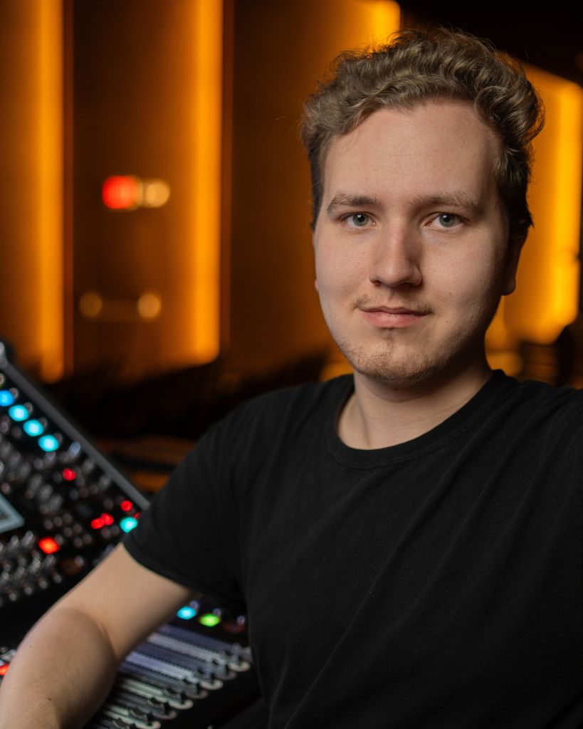 A person with dirty blonde hair wears a black t-shirt while positioned in front of music production equipment with red, blue, and green illuminated controls. Warm orange and yellow studio lighting glows in the background.