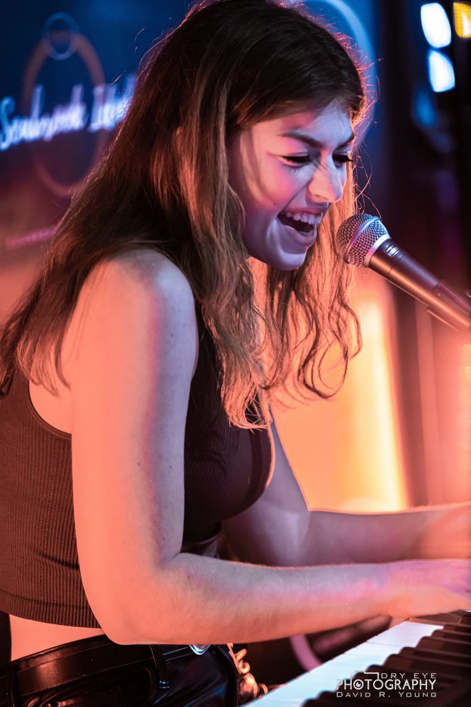 A young woman passionately singing into a microphone while playing the piano, illuminated by warm stage lighting with a vibrant and energetic atmosphere