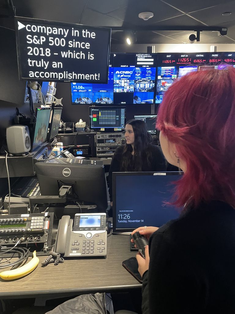Miya Menscher is seen from behind, holding a communication device while observing a control room filled with monitors, computers, and broadcasting equipment. Another individual with long dark hair is seated at a workstation, focused on the screens. A large display in the room shows text about a company being in the S&P 500 since 2018. The environment is dimly lit, illuminated primarily by the screens and equipment