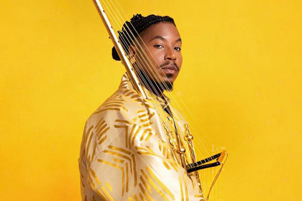 A person wearing a gold and cream patterned outfit poses against a bright yellow background, holding a stringed musical instrument with golden accents. The individual has braided hair styled neatly and looks confidently toward the camera, creating a vibrant and artistic composition.