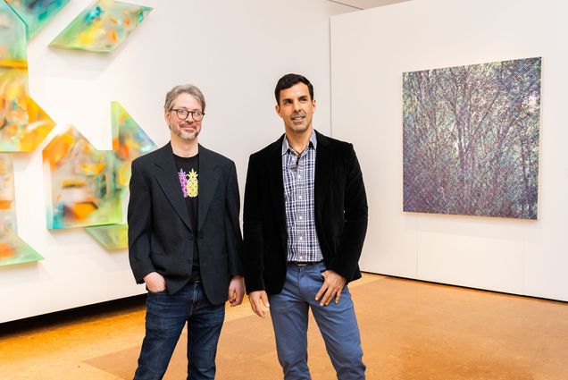 Colleagues and friends Michael Zachary (left) and Juan José Barboza-Gubo standing in BU's Faye G., Jo, and James Stone Gallery for their joint exhibition, Trasluz / Translucent. The man on the left wears glasses, a blazer, jeans, and a T-shirt with colorful pineapples. The man on the right is dressed in a plaid shirt, blue pants, and a dark velvet blazer. Behind them, on the left, is a three-dimensional geometric installation with bright, translucent colors. On the right, a square canvas displays an intricate, abstract pattern resembling a forest scene. The gallery has a clean, modern interior with bright lighting and a polished wooden floor.