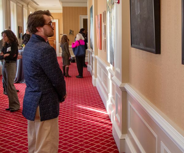 An exhibition attendee with glasses and a navy blue patterned blazer stands in a well-lit hallway, attentively observing artwork displayed on the walls. The hallway features a red patterned carpet, white trim, and other guests viewing the art in the background.