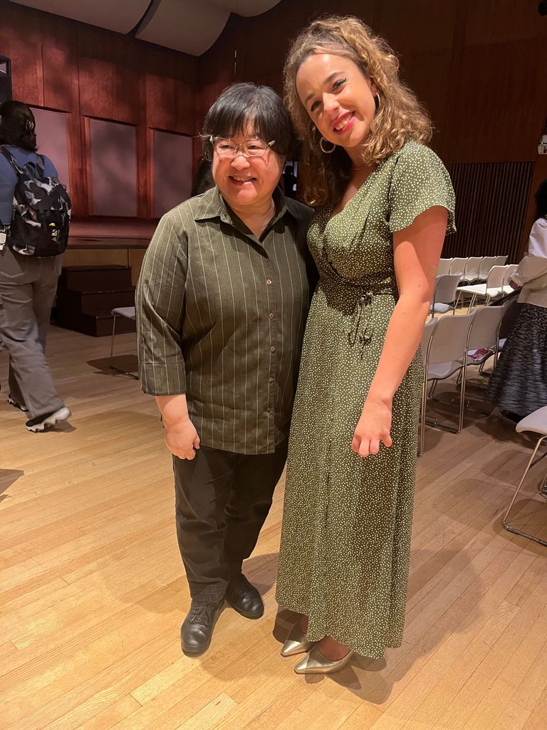 Composer Chen Yi and Boston University vocal performance major Sadie Habas (CFA'25) standing together and smiling warmly for the camera in a concert hall. On the left, Chen Yi with short black hair wearing a dark striped shirt. On the right, Sadie Habas with curly hair in a green dress with white polka dots, wearing silver heels. They are surrounded by rows of chairs and the wooden-paneled backdrop of a music performance venue.
