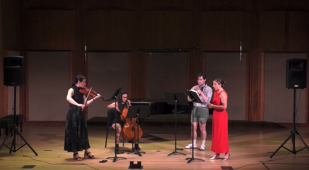 A musical ensemble performing on stage, featuring four musicians. From left to right: a violist in a long black dress, a cellist seated and playing, a clarinetist wearing a light shirt and shorts, and a soprano vocalist in a vibrant red dress. They are positioned in front of music stands, and the stage is illuminated with soft, warm lighting, creating an elegant and focused atmosphere.