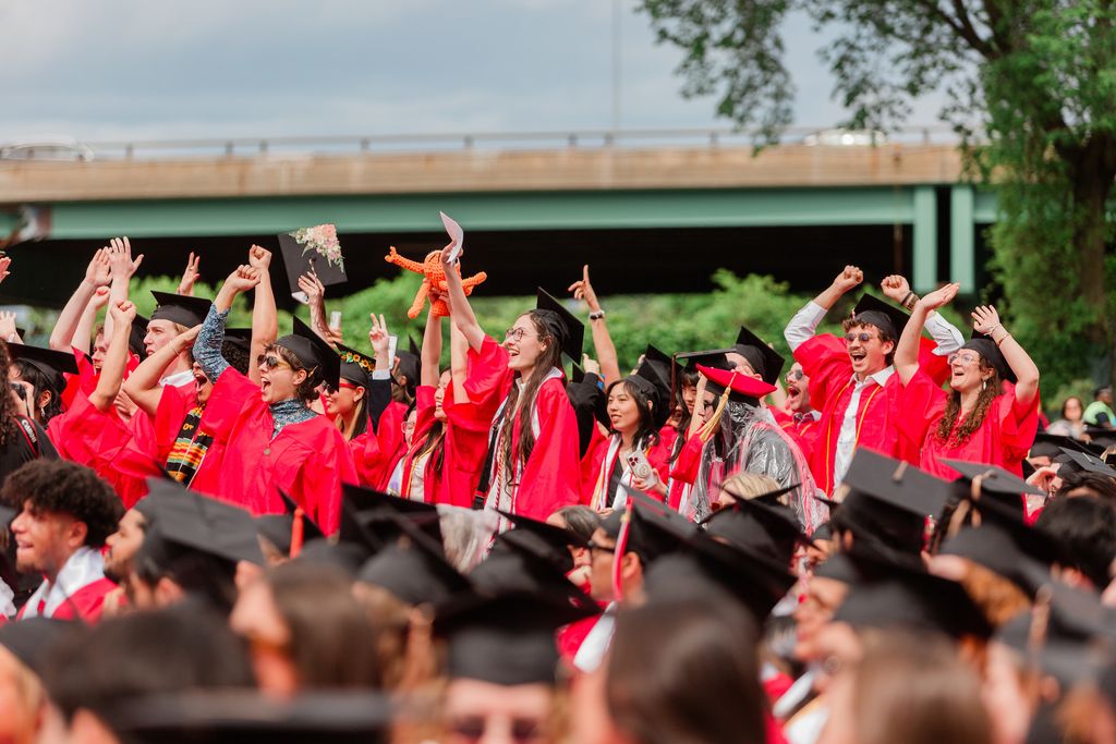 Graduating students from the Boston University Class of 2025 cheer enthusiastically in red regalia and black caps, some waving diplomas and holding celebratory items, during the outdoor Commencement ceremony.