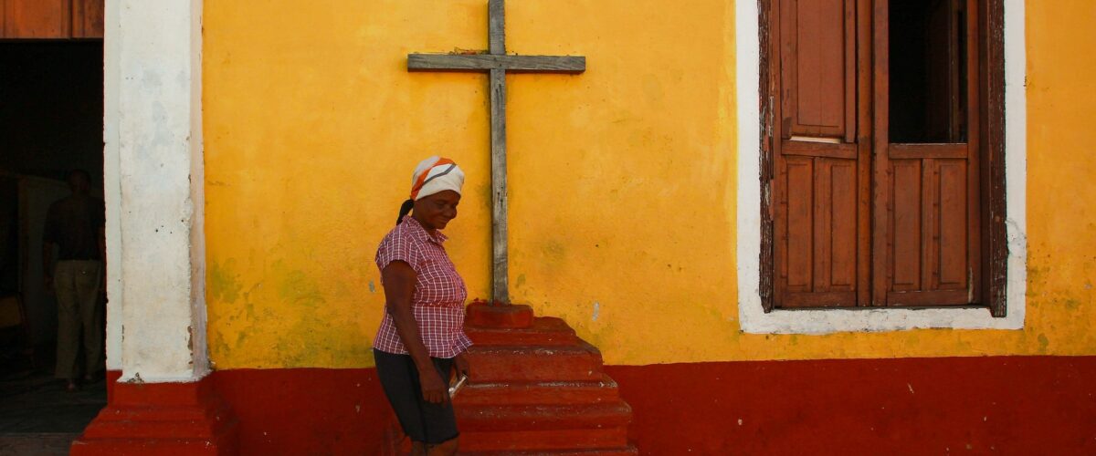 a woman walking in front of a wooden cross set up against a yellow colored wall