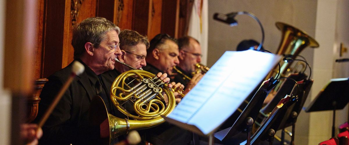 Musicians play various brass instruments. In the foreground, there is a music stand with a light and music open on it.