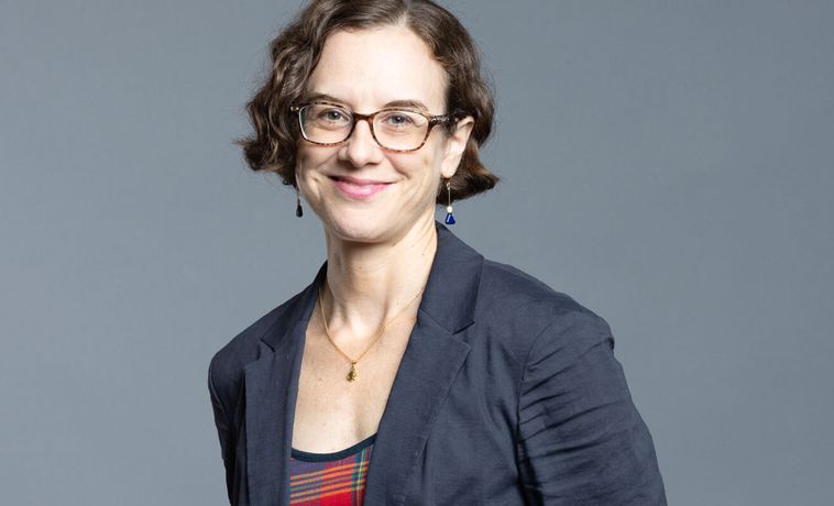 A white women with short brown hair and a gray blazer and square glasses.