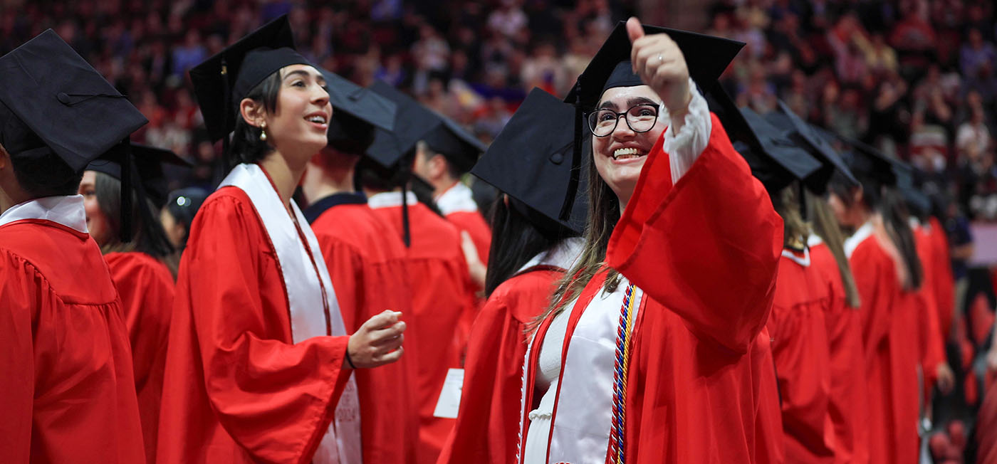 Two students wearing caps and gowns during COM Commencement ceremonies. The woman on the right is signaling thumbs-up to the crowd.