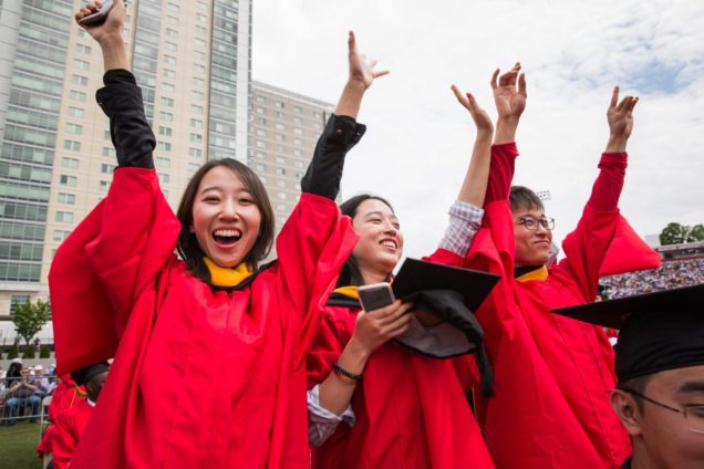 3 graduates in BU red gowns cheer with arms raised in the air.