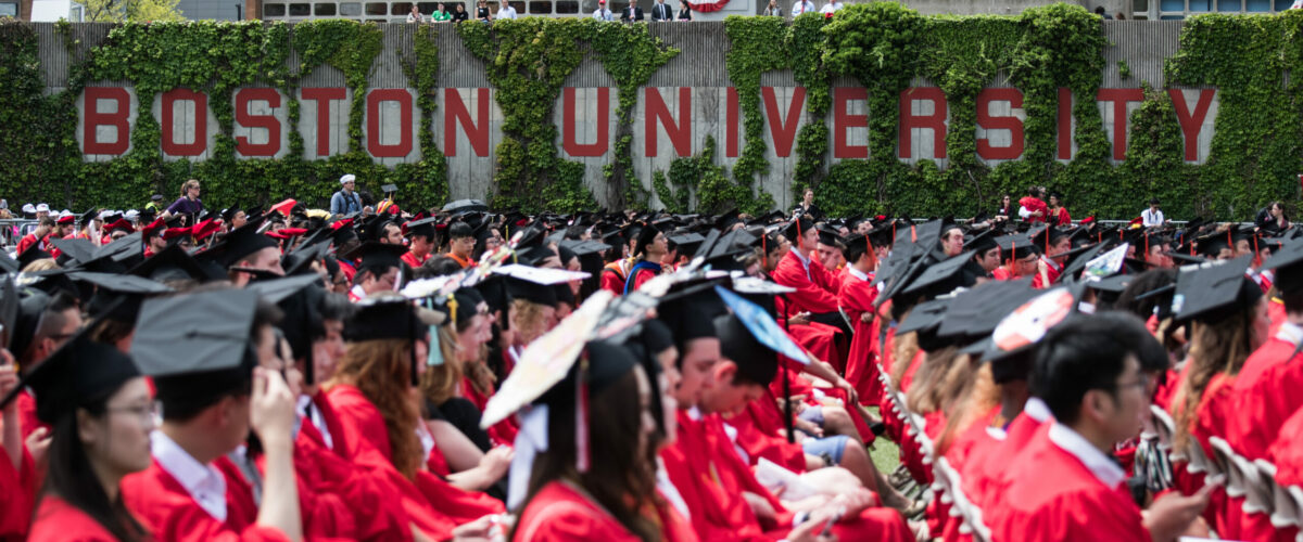 Side view of seated graduates in regalia by the Boston University sign.