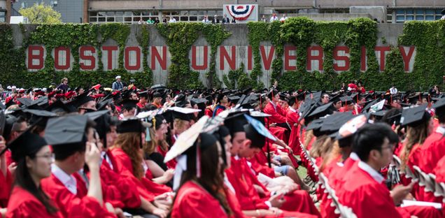 Side view of seated graduates in regalia by the Boston University sign.