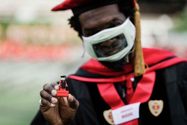 An advanced degree graduate holds up his LEGO BU Graduate figure for the camera. The LEGO graduate is wearing red with black stole and black mortarboard cap.