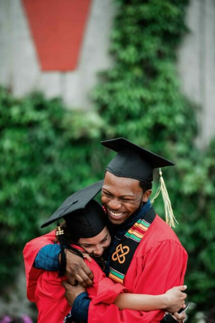 2 graduating students wearing cap and gown embrace in a big hug following Commencement.